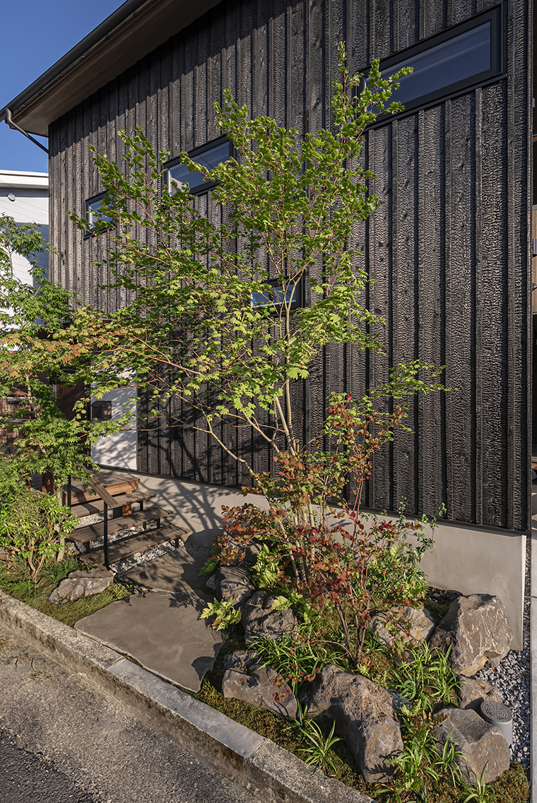 引き締まった黒い天龍焼杉の建物外観に合わせて、鉄平石の石敷きとウッドデッキ階段でシックにまとめ、雑木や苔を配したアプローチ。